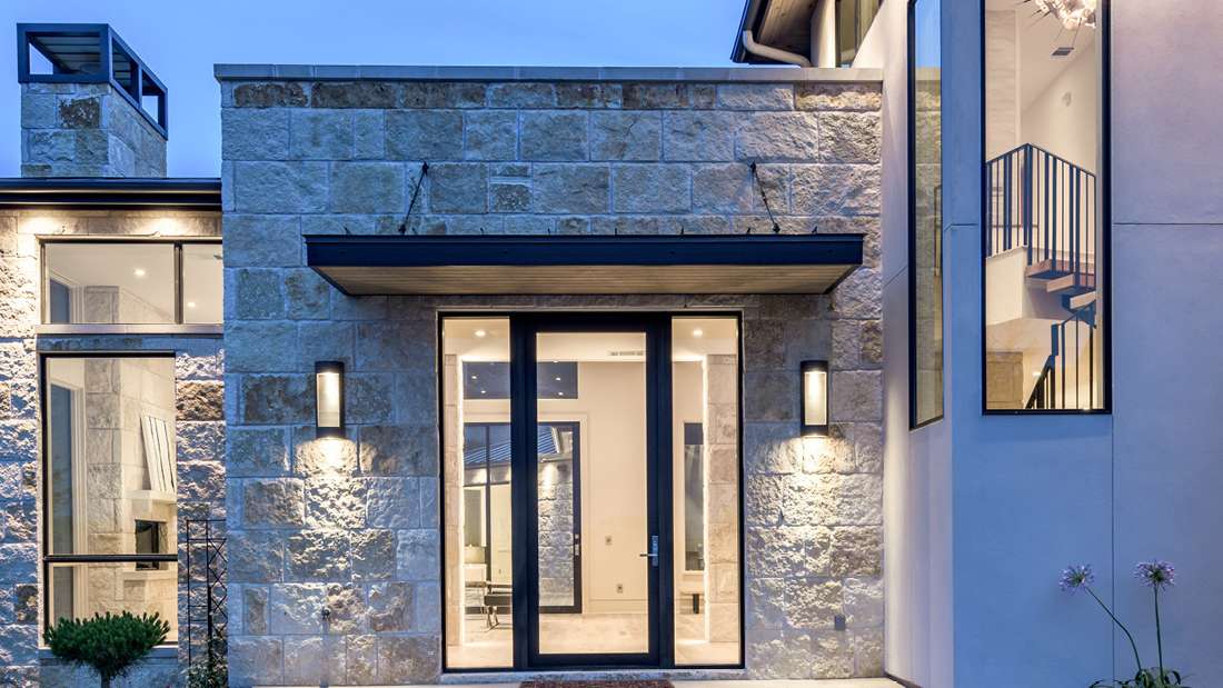 Contemporary home entrance with natural stone facade, black-framed glass double doors, modern wall sconces, and large vertical window showcasing staircase inside.