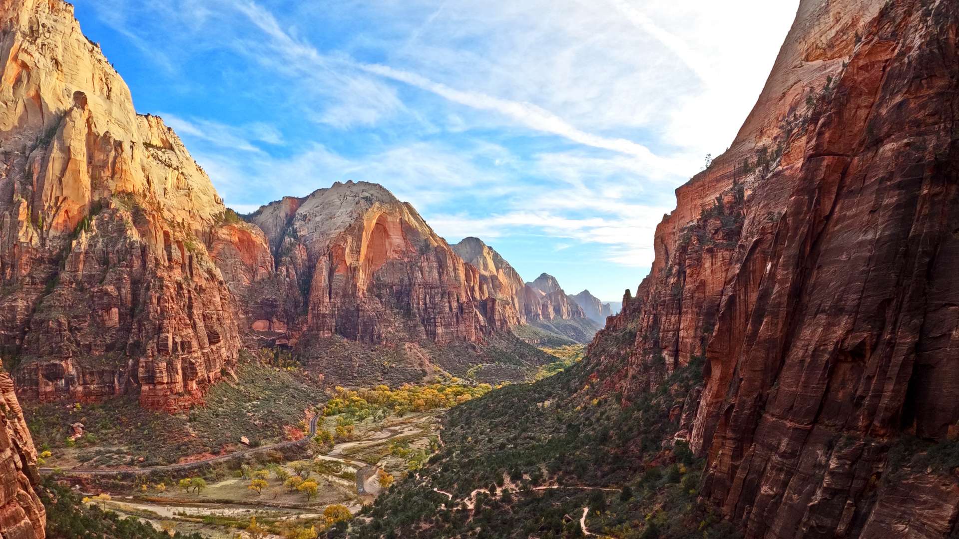   Panoramic view of a canyon with towering red cliffs, winding valley trails, and a bright blue sky.