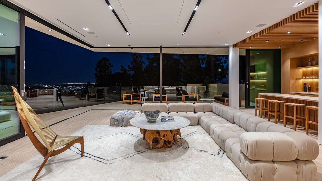 Luxury open-concept living room with panoramic city views at night, floor-to-ceiling glass doors, modular beige sectional sofa, round stone coffee table with natural wood base, and modern bar area with wooden stools.