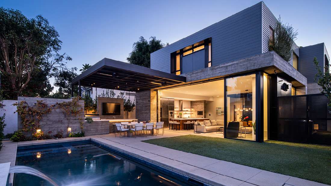 Luxury modern home exterior at dusk with floor-to-ceiling glass walls, open-concept living area, covered outdoor dining space featuring built-in TV and lighting, and rectangular swimming pool with water feature.