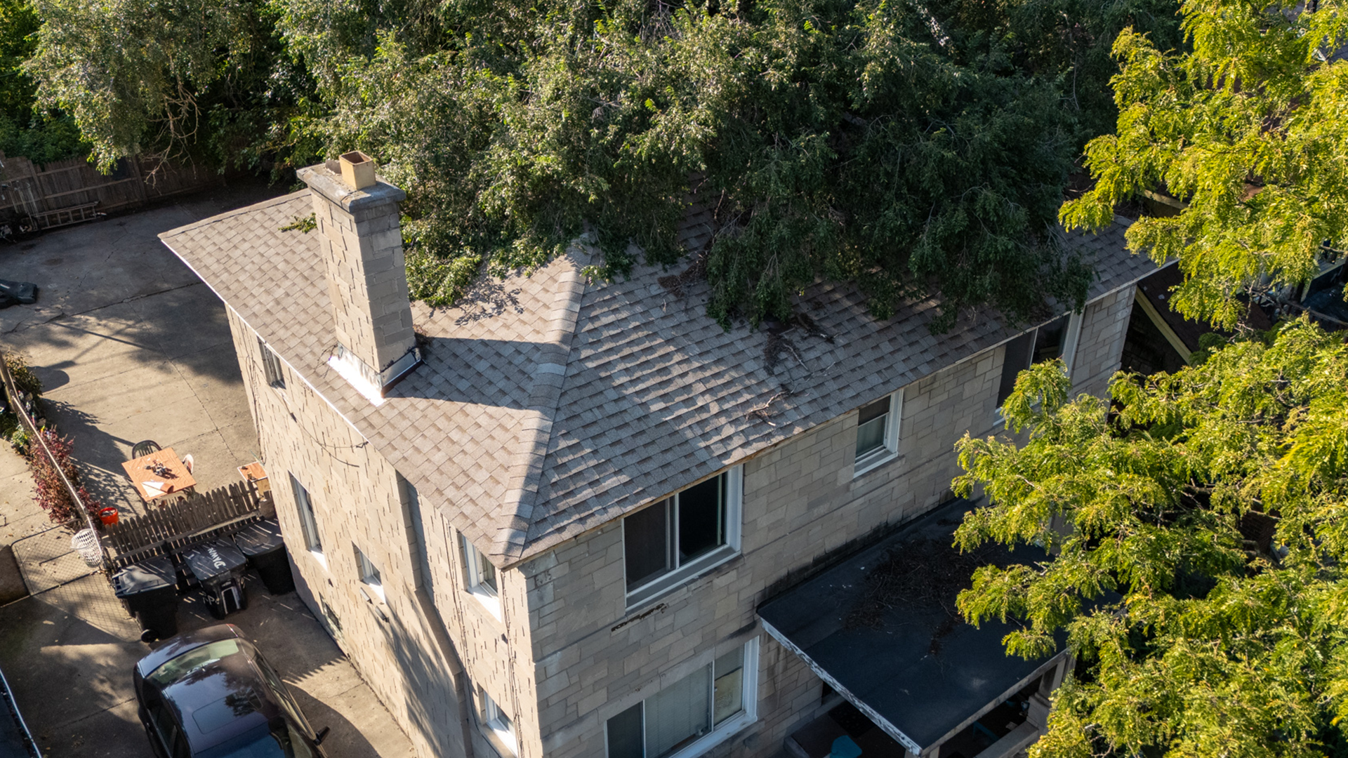 Tannish-grey brick home with old, damaged shingles and trees excessively grown over the rooftop.