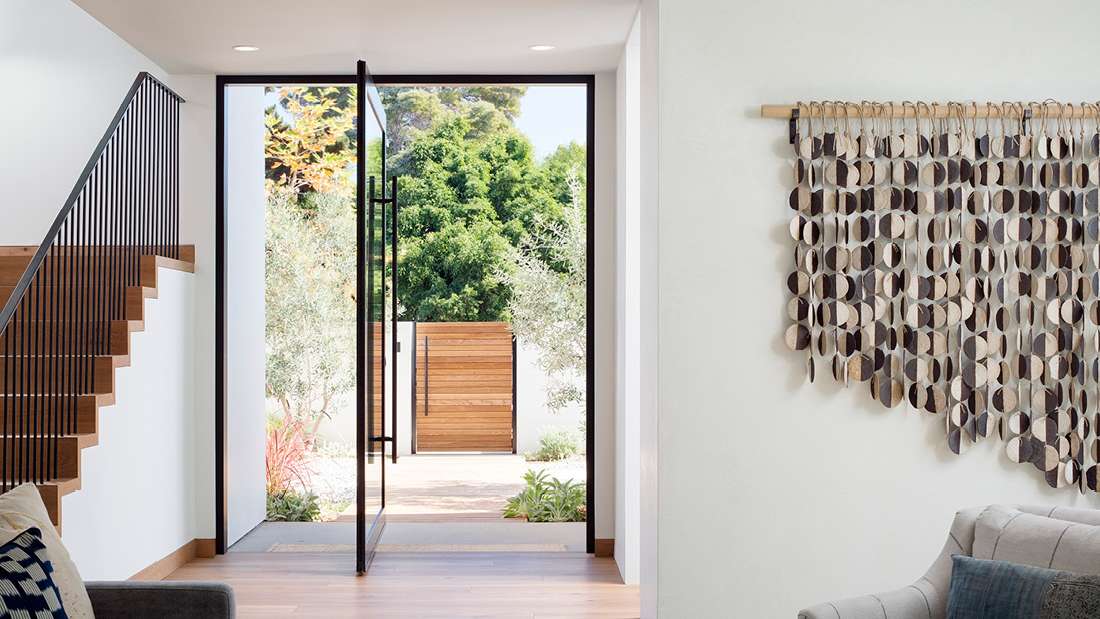 Modern entryway with pivot glass door framed in black metal, wooden staircase with slim vertical railings, light wood flooring, and outdoor view featuring wooden gate and lush greenery.