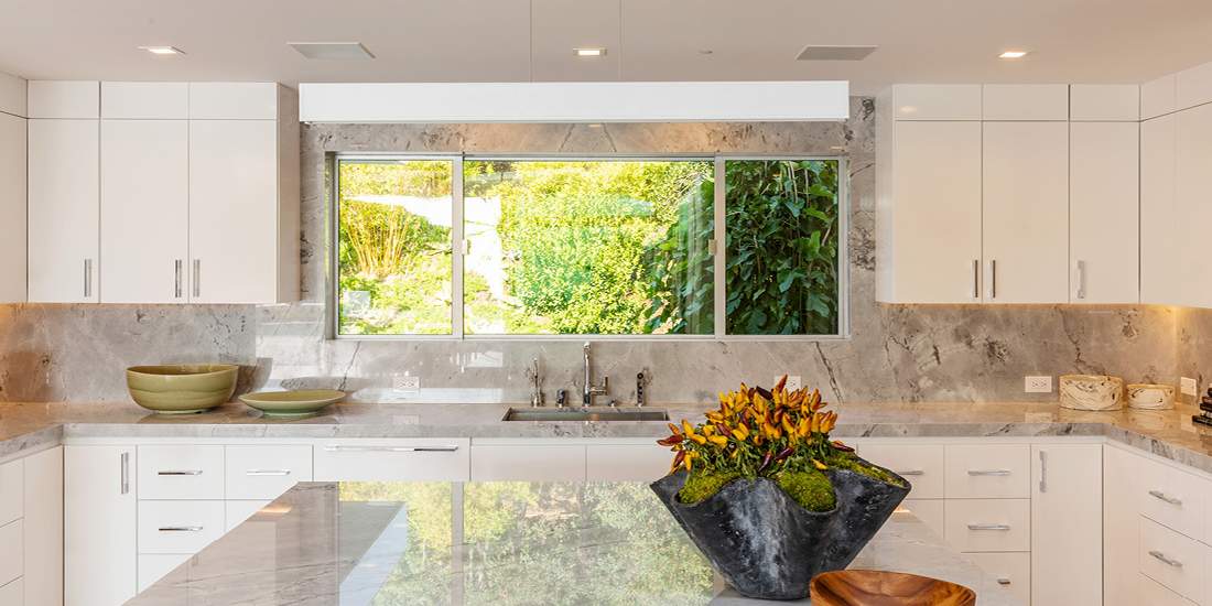 Luxury modern kitchen with white cabinetry, marble countertops and backsplash, large center island with polished stone surface, and wide window overlooking lush green garden.