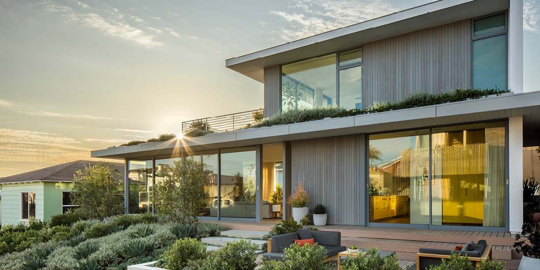 Modern two-story home with vertical wood siding, large glass windows, rooftop greenery, and landscaped front yard featuring succulents and outdoor seating at sunset.