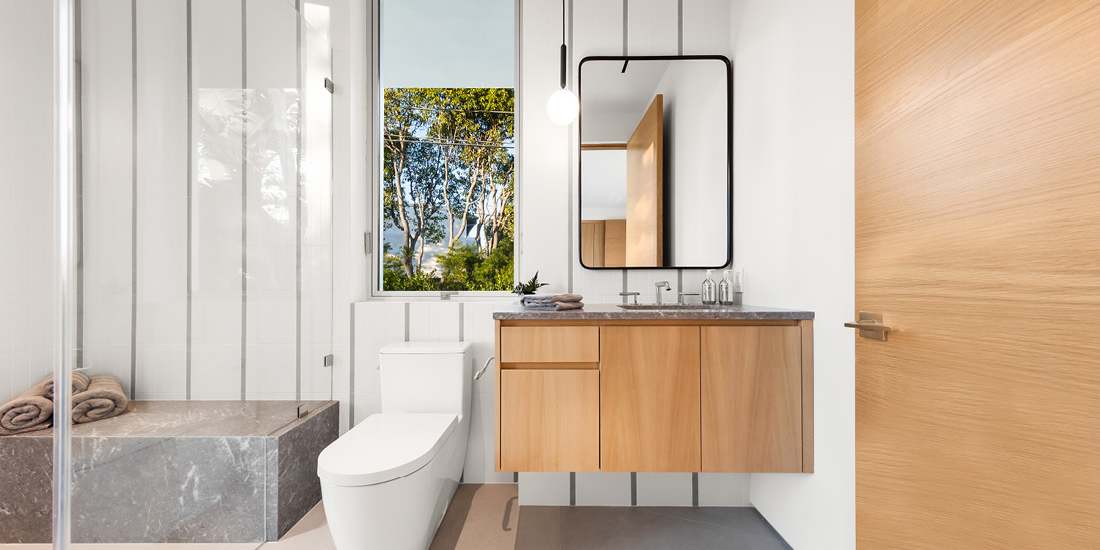 Modern bathroom with frameless glass shower enclosure, gray stone bench, white toilet, floating wood vanity with dark stone countertop, black-framed mirror, and pendant light, featuring a large window with outdoor greenery view.