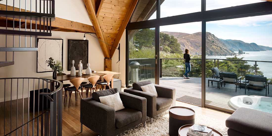 Rustic modern living and dining space with exposed wooden beams, spiral staircase, cozy seating area, and floor-to-ceiling glass doors opening to a deck with ocean and cliff views.