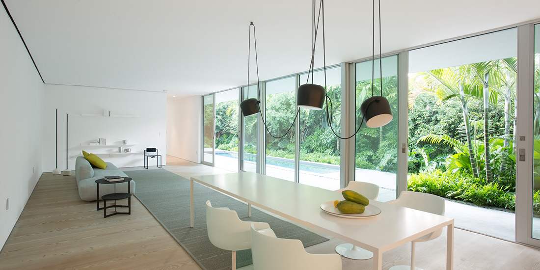 Minimalist open-concept living and dining area with floor-to-ceiling sliding glass doors overlooking lush tropical garden, featuring white dining table with modern chairs, black pendant lights, light wood flooring, and sleek gray sofa.