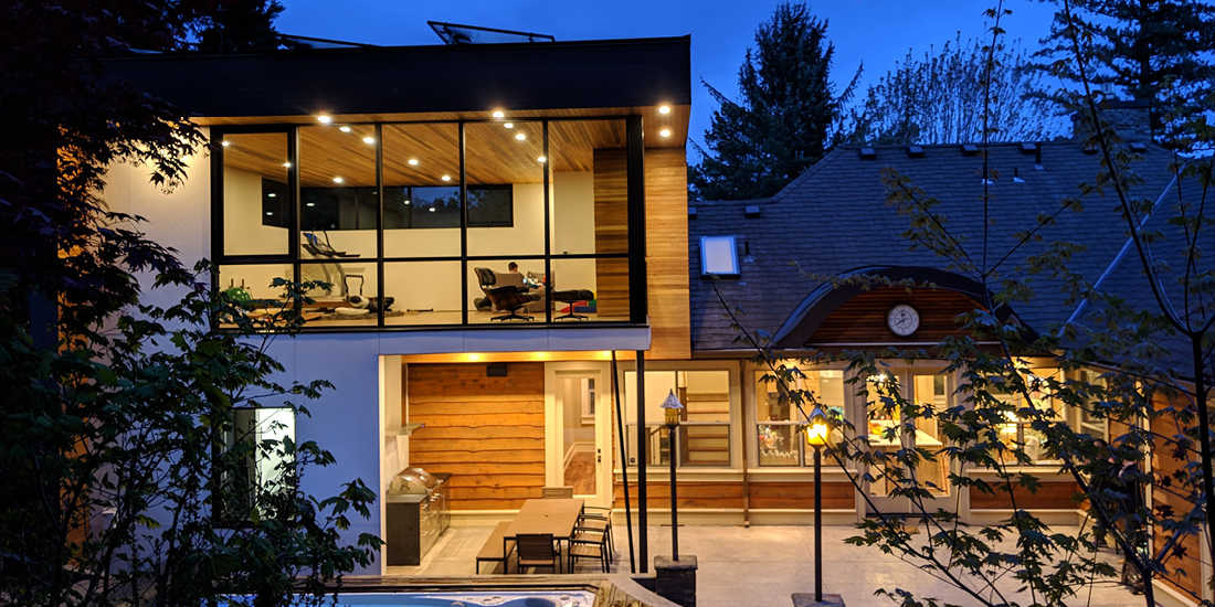 Contemporary home exterior at dusk featuring two-story glass walls, wood-paneled ceiling with recessed lighting, outdoor dining area with built-in grill, and elevated hot tub surrounded by trees.