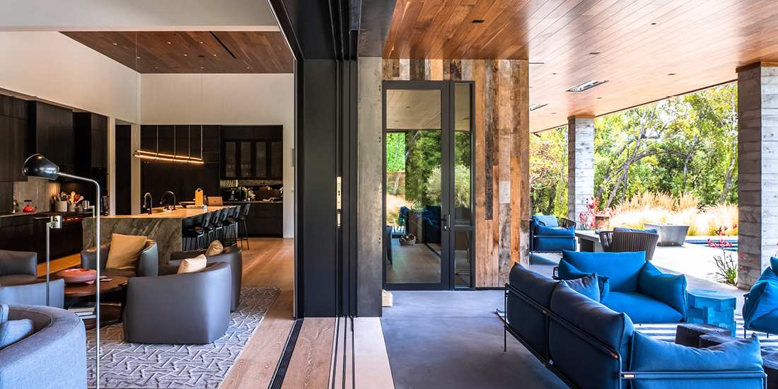 Luxury open-concept living and kitchen area with natural wood ceiling, black cabinetry, marble island with barstools, and folding glass doors opening to a covered patio featuring blue lounge chairs and outdoor greenery.