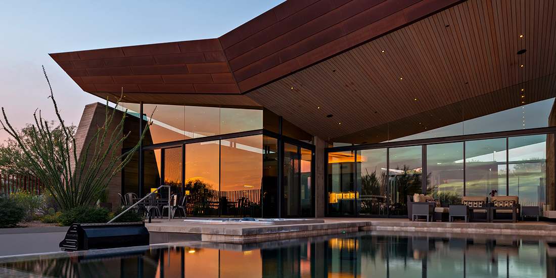 Contemporary desert home with angular roof design, floor-to-ceiling glass walls, outdoor seating area, and infinity pool reflecting a vibrant sunset.