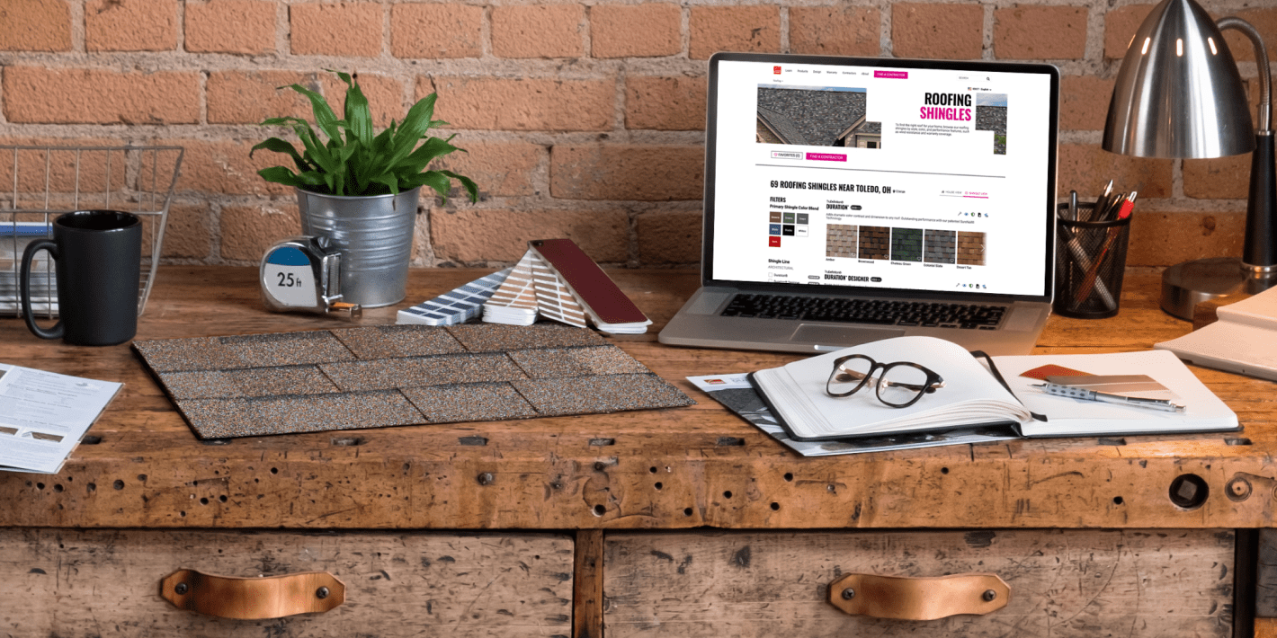 Wooden desk with Owens Corning Roofing brochures open to view shingles, a computer with the Owens Corning website displaying the Pick Your Shingles page, a black coffee mug, a tape measure, and other miscellaneous office equipment.