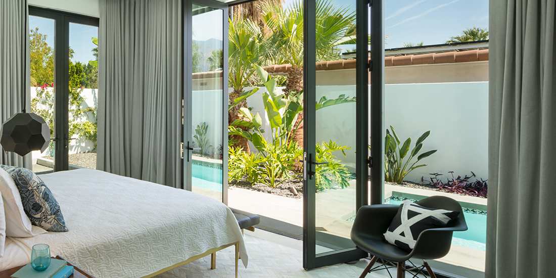 Modern bedroom with floor-to-ceiling glass doors opening to a private patio with pool, gray curtains, white bedding, and black accent chair.