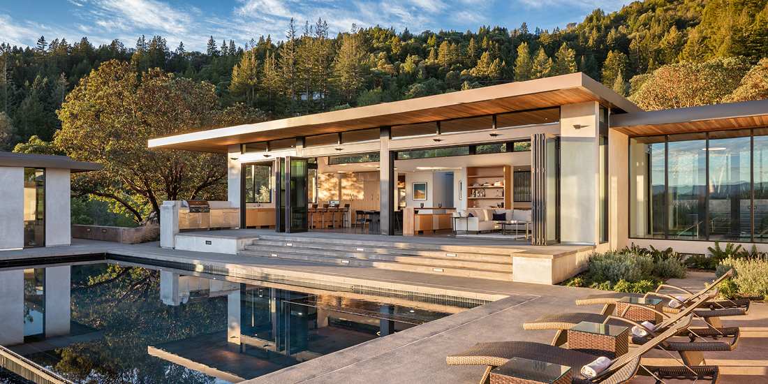Luxury modern hillside home with flat roof design, floor-to-ceiling glass walls, open-concept living area, and outdoor terrace featuring infinity-edge pool and lounge chairs overlooking forested mountains.