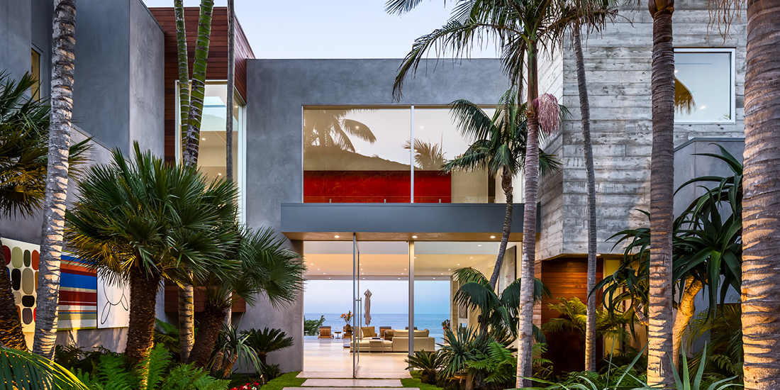 Luxury modern coastal home exterior with minimalist concrete walls, glass balcony railings, vertical wooden slats, and floor-to-ceiling glass doors framed by tropical landscaping featuring palm trees and ocean views.