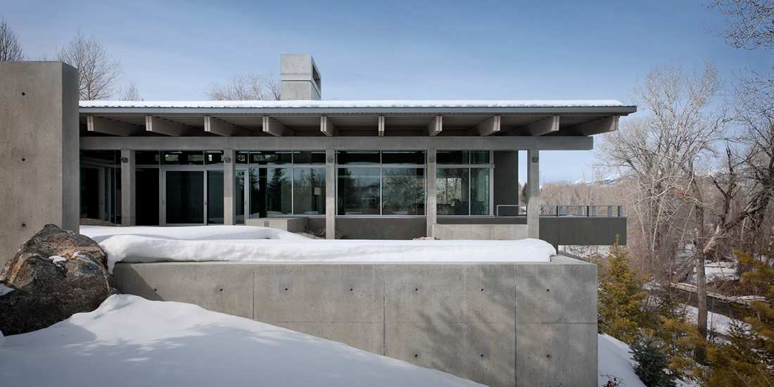 Modern mountain home with concrete exterior walls, flat roof design, and large glass windows surrounded by snow-covered landscape and bare winter trees.