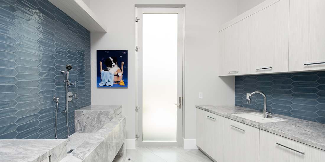 Modern laundry and pet wash room with blue textured tile backsplash, marble countertops, built-in white cabinetry, and frosted glass door providing natural light.