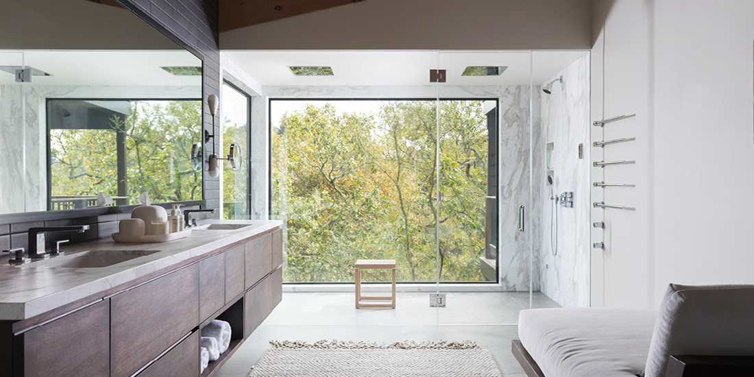 Luxury bathroom with double-sink vanity featuring dark wood cabinetry and marble countertop, large wall mirror, glass-enclosed shower with rainfall fixtures, and floor-to-ceiling windows overlooking lush green trees.