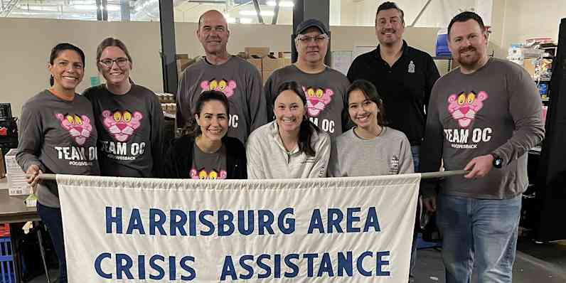 A group of people stand wearing matching shirts smiling holding a sign that reads "harrisburg area crisis assistance"