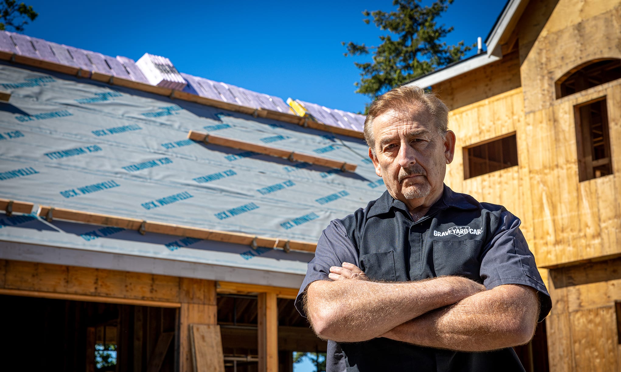 Mark Worman wearing a grey Graveyard Carz shirt standing in front of his partially finished home showing Titanium X30 underlayment installed on the roof deck.