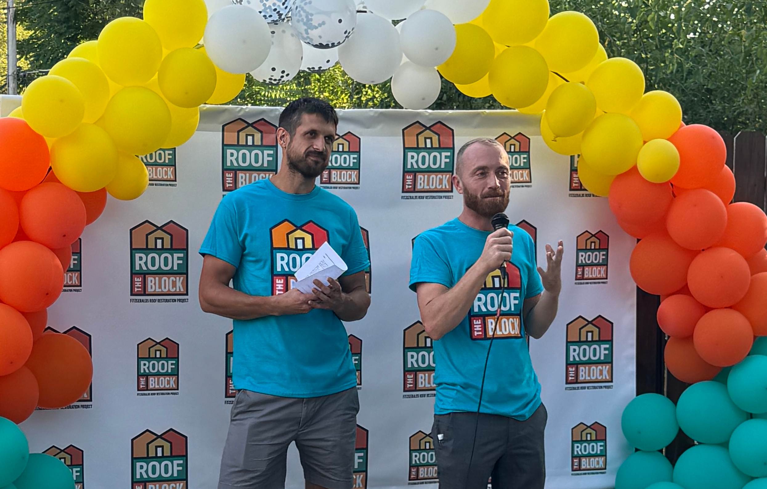 Keith and Evan from HGTV Bargain Block stand in front of a balloon arch while delivering their speech to the roof recipients. 