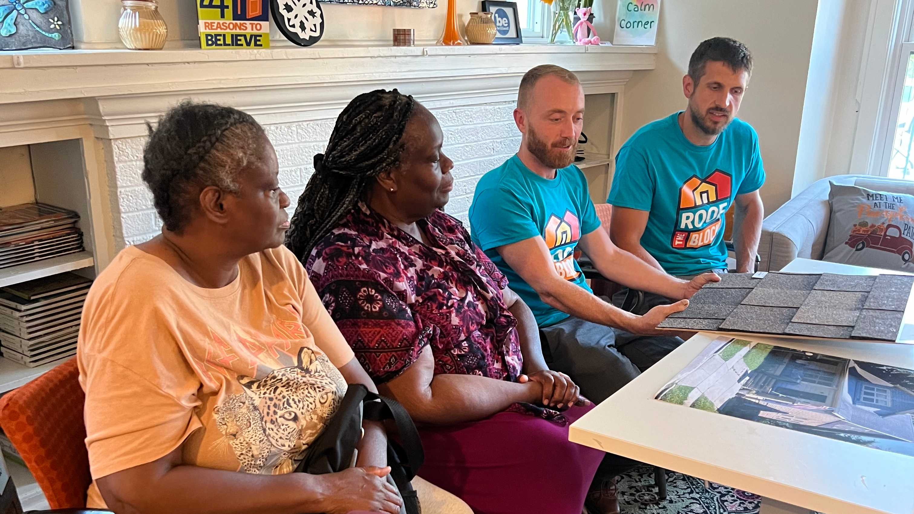 Two homeowners sitting at a white table with Keith and Evan looking at roof shingle sample boards and a large photo of their home during the design consultation.
