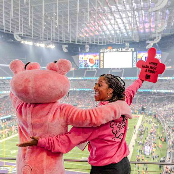 La mascota de la Pantera Rosa y una mujer con una sudadera rosa con la cara de la Pantera Rosa y extendiendo un dedo de espuma rosa que dice "Más que solo un techo" frente al campo del Allegiant Stadium.