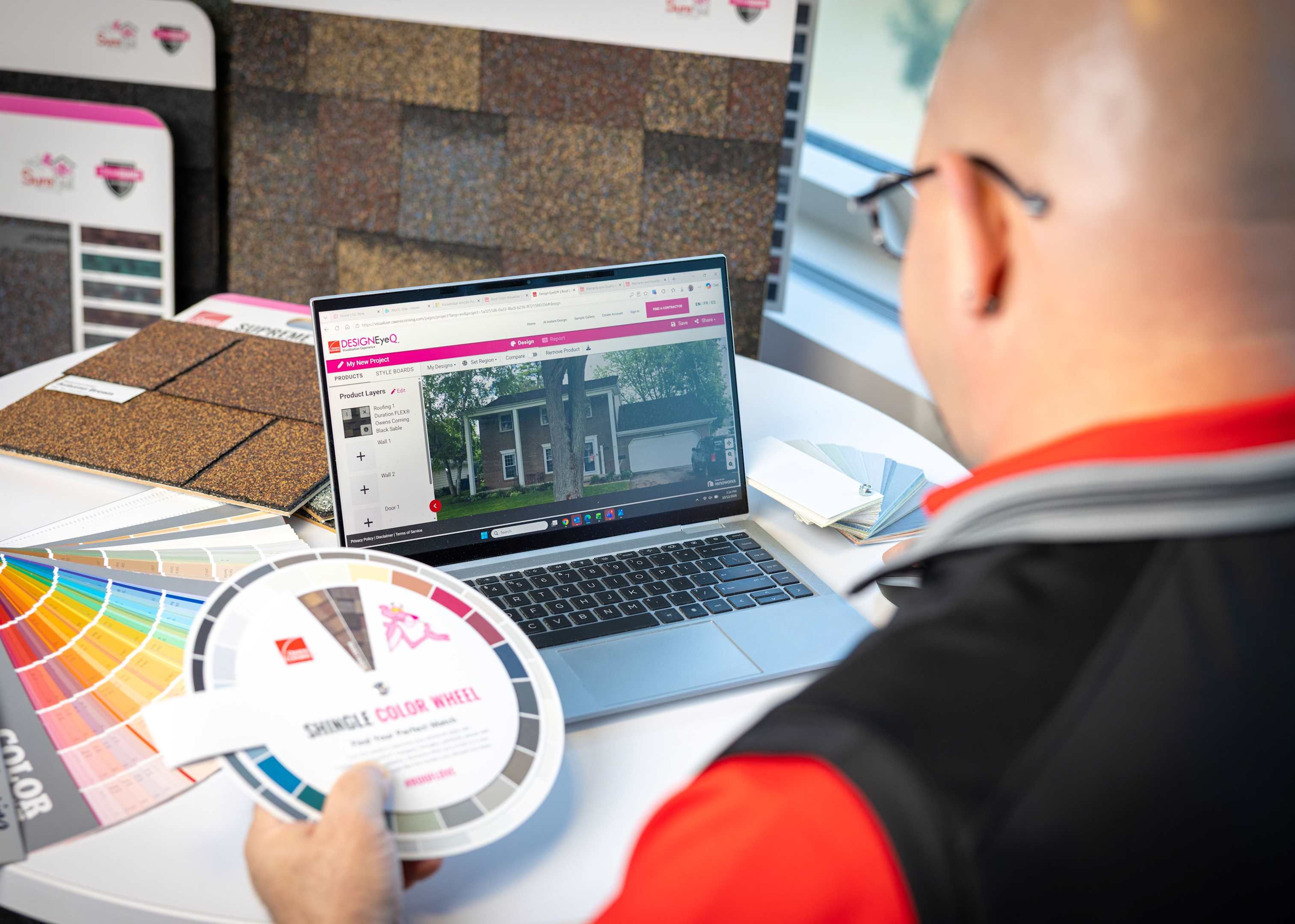 Male color consultant with headset on his laptop with 2 monitors with Design EyeQ and shingles page open. Paint swatch book open on desk,