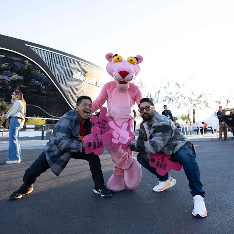 Pink Panther mascot and woman wearing a pink sweatshirt with the Pink Panther face and holding out a pink foam finger that says "More than Just a Roof" facing the field of Allegiant Stadium.