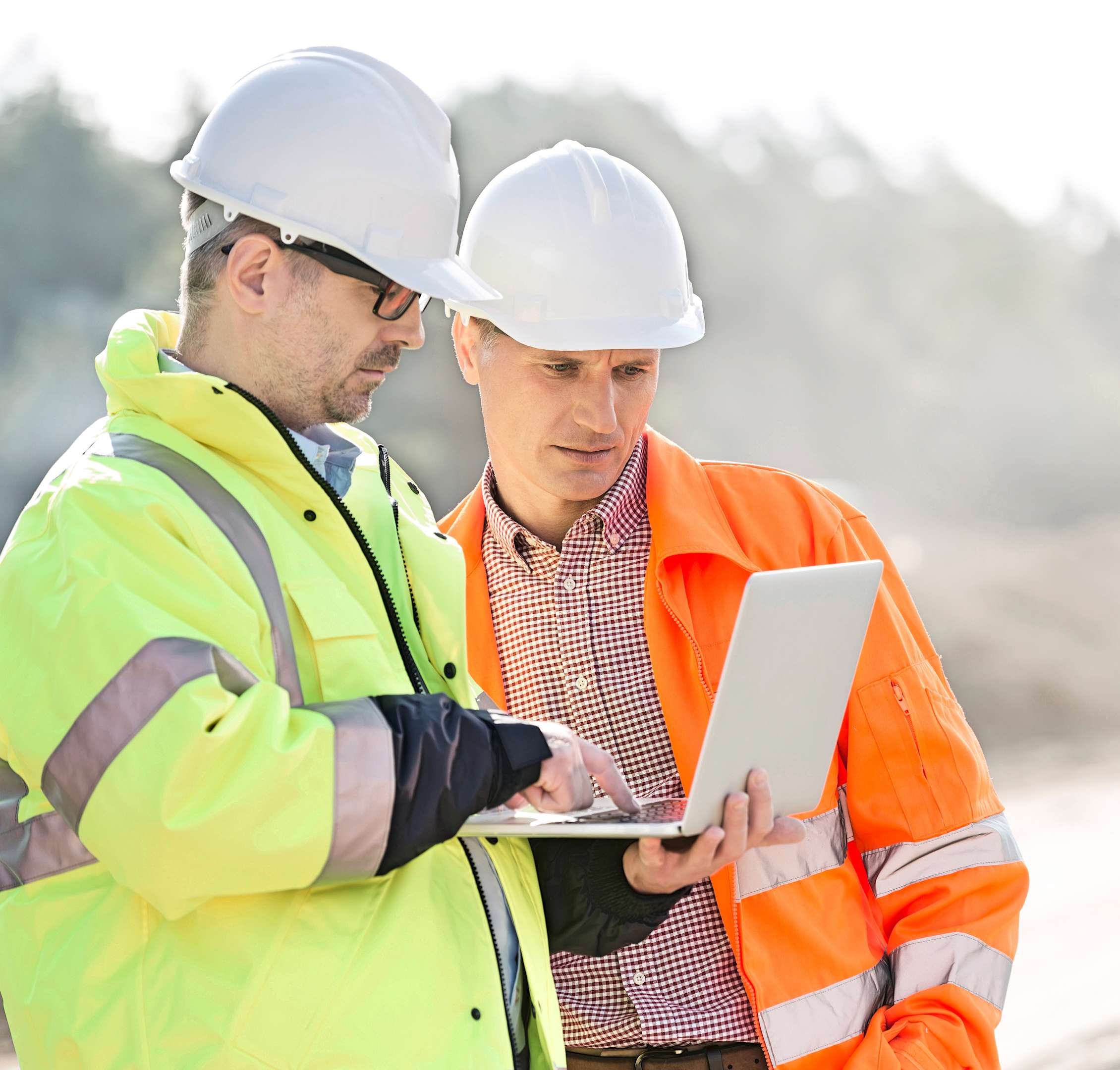 Two people wearing reflective jackets and helmets are standing outside looking at a computer.