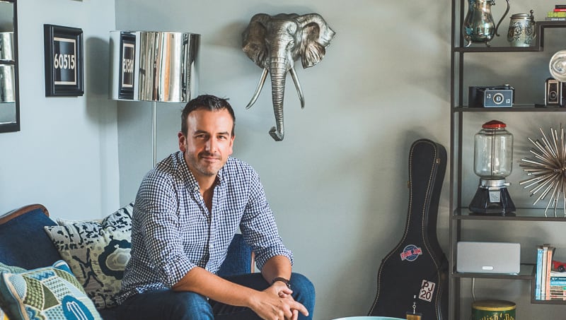 Chad Esslinger sitting on a blue couch in his livingroom with eccentric home decor, including a chrome elephant head on the wall