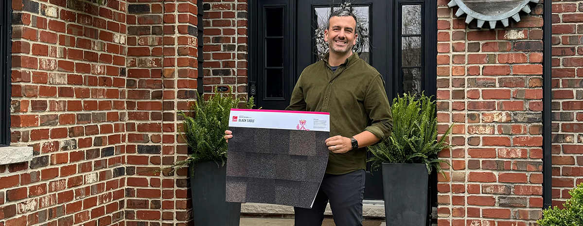 Chad Esslinger standing in front of his red brick home and black metal door with windows and sidelites, holding an Owens Corning Black Sable printed shingle poster.