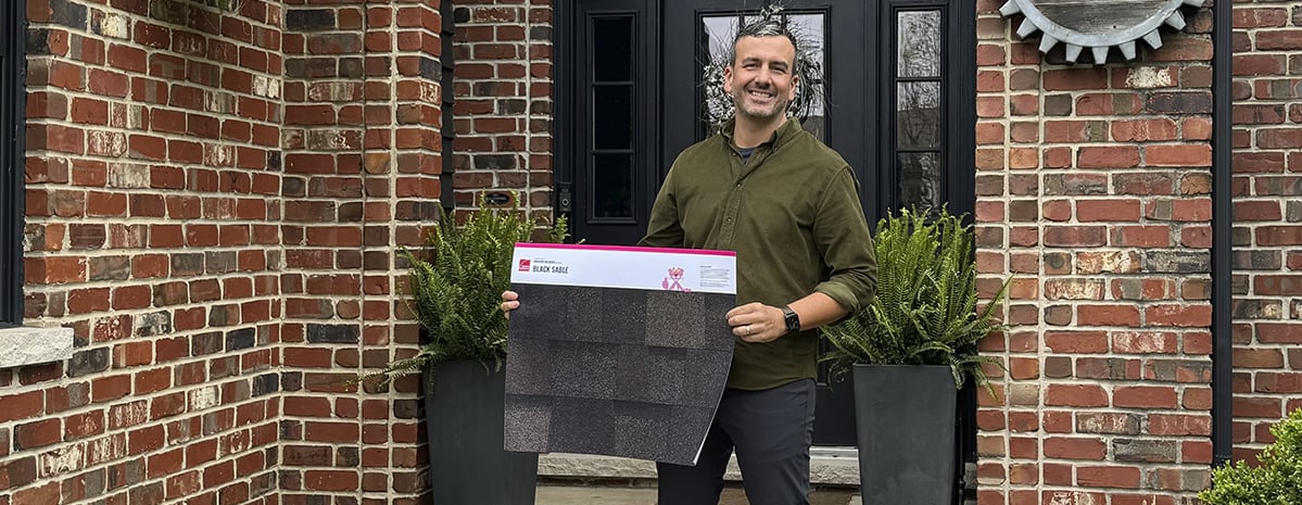 Chad Esslinger standing in front of his red brick home and black metal door with windows and sidelites, holding an Owens Corning Black Sable printed shingle poster.