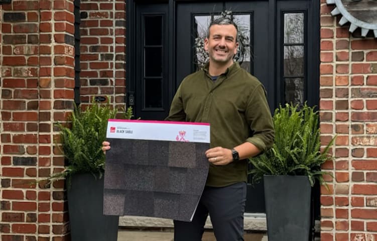 Chad Esslinger standing in front of his brick home with black door, holding a Black Sable shingle color poster.