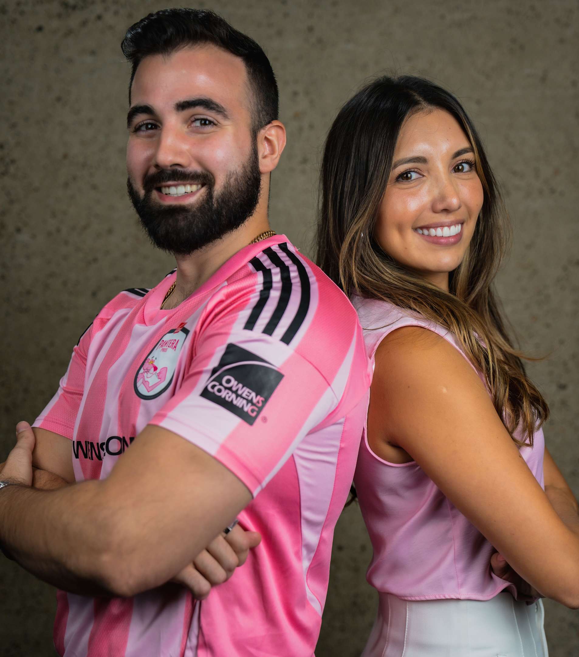 Headshot of a smiling young woman with long brown hair, parted in the middle, wearing a light pink v-neck top with a small floral print. She is positioned slightly off-center, and the background, slightly blurred, shows trees, shrubs, and a paved area, suggesting an outdoor setting, possibly a patio. She is wearing small earrings.