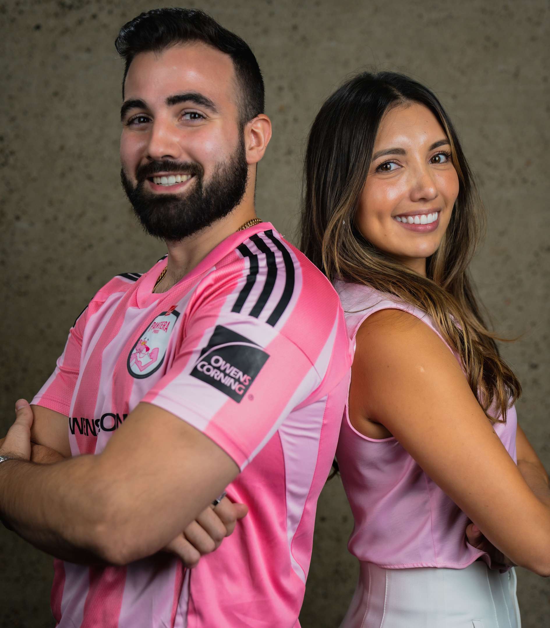 Headshot of a man with short dark hair and a beard, wearing a bright pink polo shirt with a small "Owens Corning" logo on the right side of the chest. He is looking directly at the camera with a neutral expression. The background is slightly out of focus and appears to be an outdoor setting with greenery and part of a building visible.
