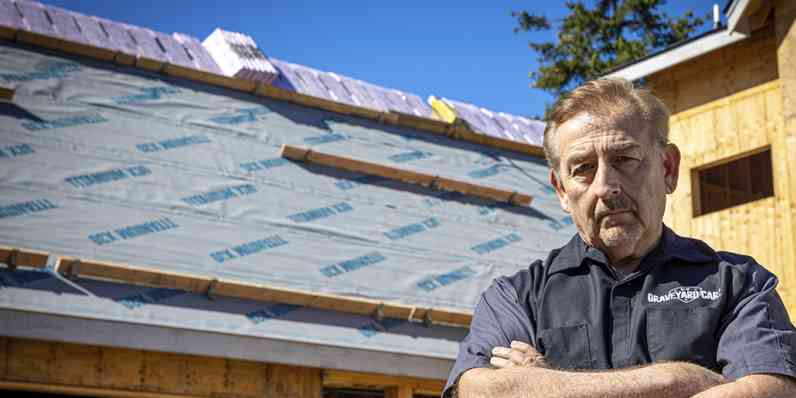 Mark Worman standing with his arms crossed in front of his in-construction dream home with underlayment spread across the roof