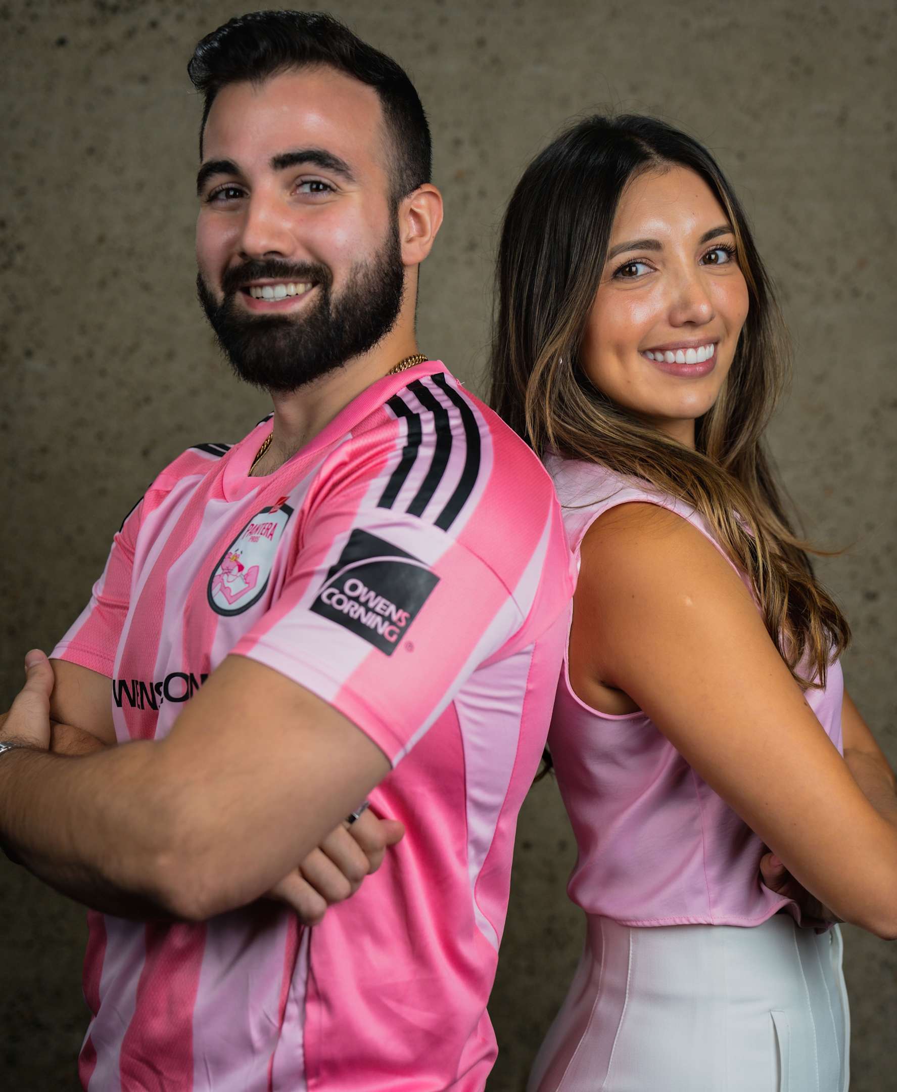 Primer plano de un hombre con cabello corto y oscuro, barba y vestido con un polo rosa brillante con un pequeño logo de "Owens Corning" en el lado derecho del pecho. Mira directamente a la cámara con expresión neutra. El fondo está ligeramente desenfocado y parece ser un exterior con vegetación y parte de un edificio visible.