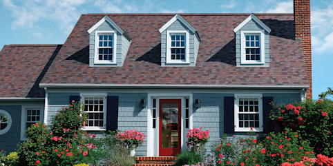 Blue grey house with Owens Corning Duration Designer shingles in Merlot, surrounded by a blue sky and shrubbery with spring flowers.