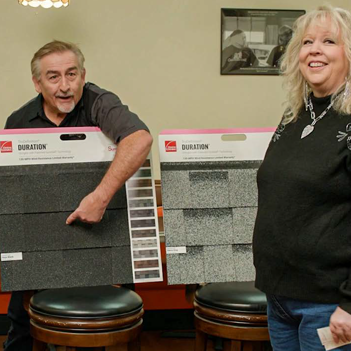 Mark Worman standing behind a shingle sample board pointing to the Black Onyx color with wife Suzanne off to the right to 
