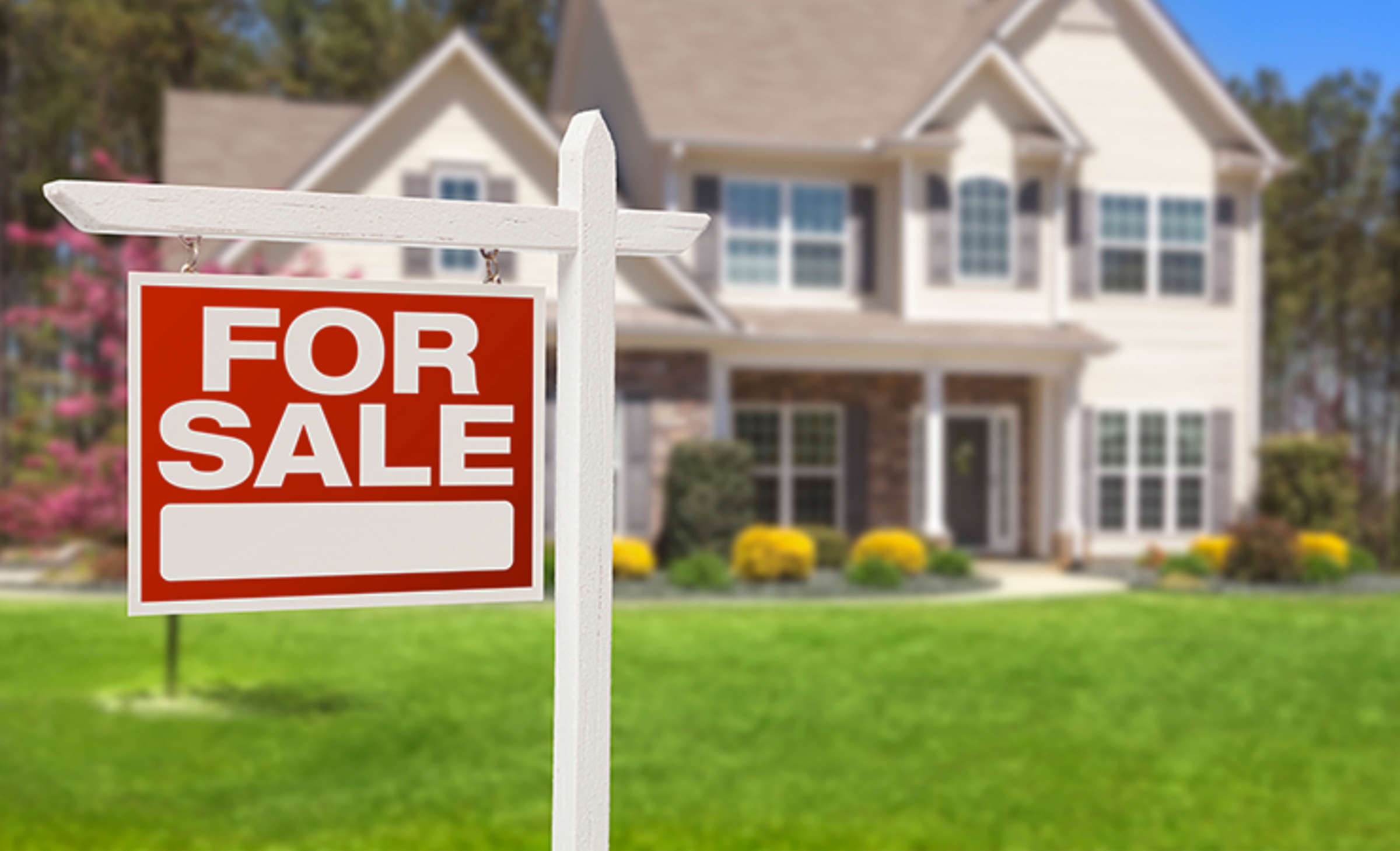Tan house with bright green lawn in the background with red "For Sale" sign in the foreground