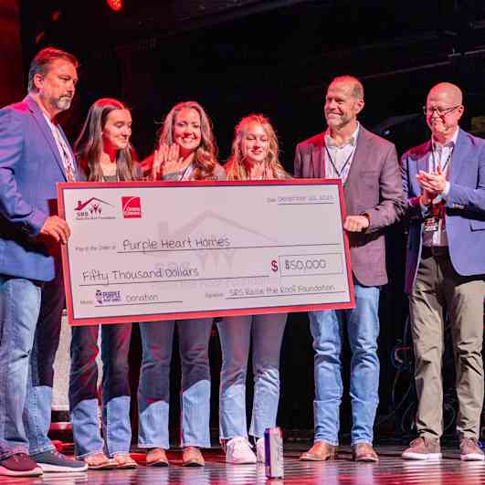 Seis personas de pie en un escenario sosteniendo un cheque de caridad de gran tamaño por valor de 50.000 dólares para Purple Heart Homes.