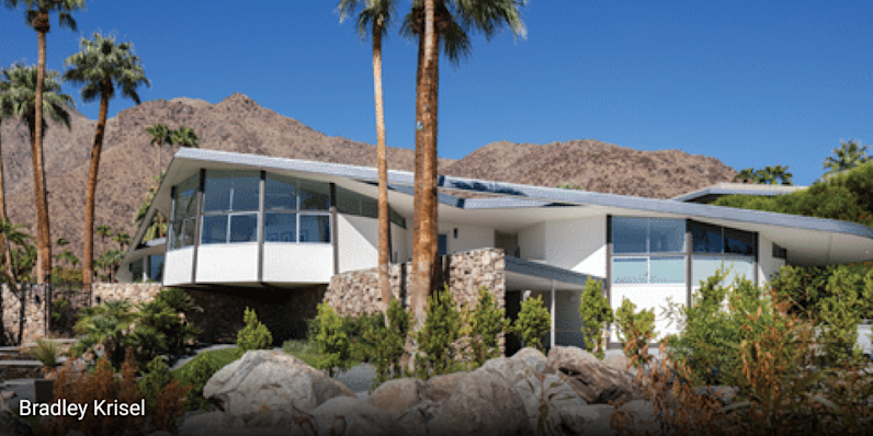 Mid-century modern home in Palm Springs, California featuring dramatic angular rooflines, cantilevered design, large glass windows, and stone exterior walls, surrounded by palm trees and desert landscaping with rocky terrain.