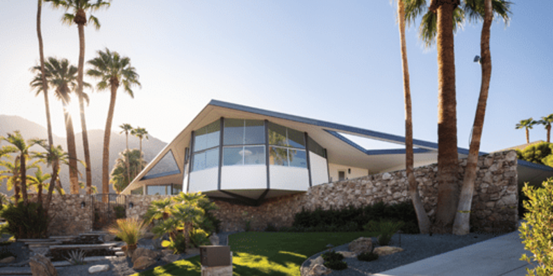 Mid-century modern home in Palm Springs, California featuring dramatic angular rooflines, cantilevered design, large glass windows, and stone exterior walls, surrounded by palm trees and desert landscaping under bright sunlight.
