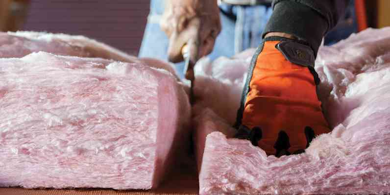 A contractor cuts a slab of pink wall insulation with a box cutter while wearing orange gloves