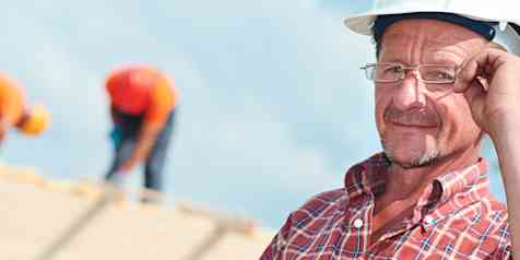 Close up of man wearing a white hard hat, adjusting glasses on his face, with blurred roofers on top of roof in background.