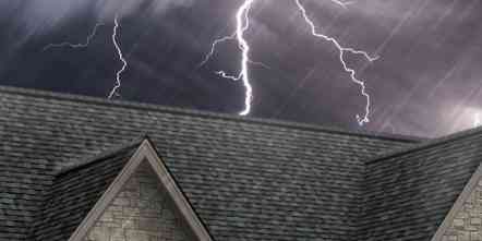 Photo of the top of a roof with gray shingles set against a dark, stormy sky with lightning