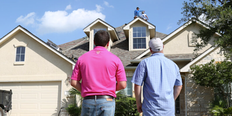 Photo of a contractor and homeowner looking at a house while roofers are working on installing shingles