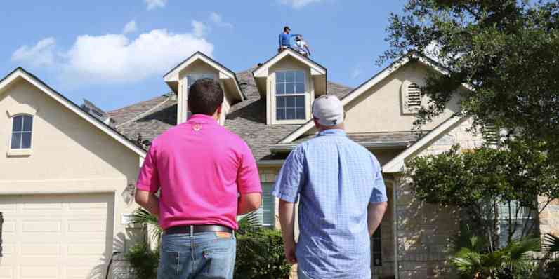 Two people in a driveway looking at an installer working on a roof