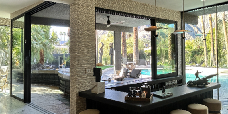 Modern indoor-outdoor living space with textured stone walls, large sliding glass doors opening to a poolside patio, black island bar with round stools, pendant lights, and landscaped garden visible through floor-to-ceiling windows.