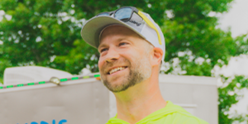 Person in a bright yellow shirt and cap standing outdoors near a trailer with trees in the background.